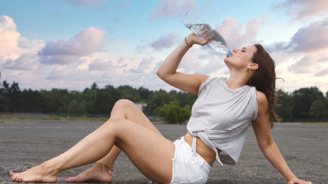 Young woman in shorts sitting on pavement and drinking wate