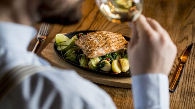 Young man eating salmon fillet with gratinated potatoes, leek and spinach in the restaurant with glass of white wine