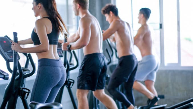 Beautiful sport women exercise with elliptical machine among the other men in the gym with day light.