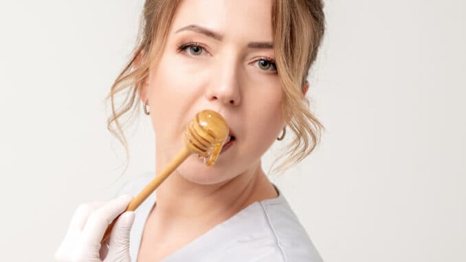 Portrait of beautiful young caucasian woman eating honey with wooden spoon on white background