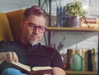 Older white man in glasses reading book at home sitting in armchair in cosy room.