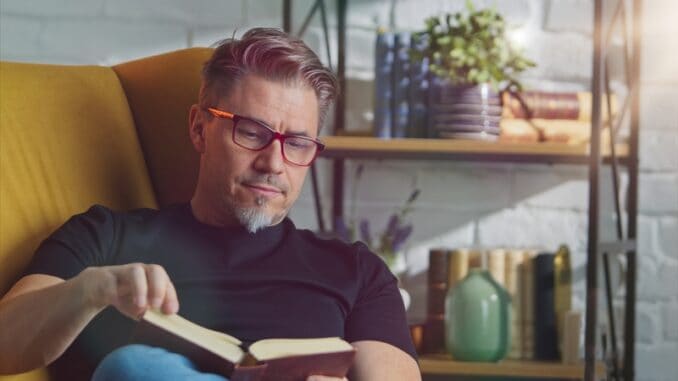 Older white man in glasses reading book at home sitting in armchair in cosy room.