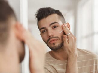 Portrait of young man applying face cream while looking in mirror during morning skincare routine