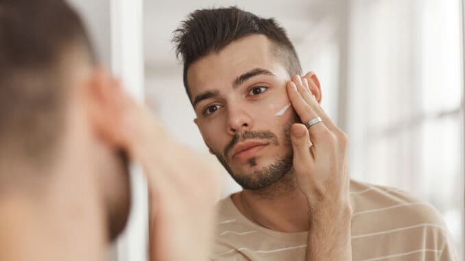 dreamstime_s_213434230 Portrait of young man applying face cream while looking in mirror during morning skincare routine
