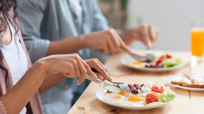 Unrecognizable couple using fork and knife while eating tasty breakfast in kitchen