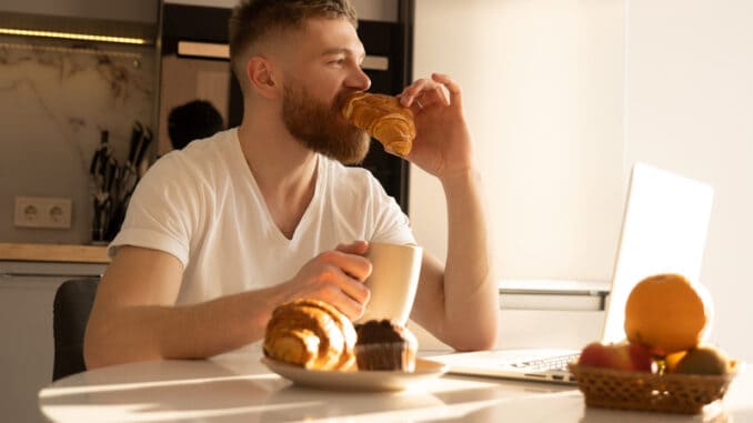 Young man eating croissant and drinking tea or coffee on breakfast