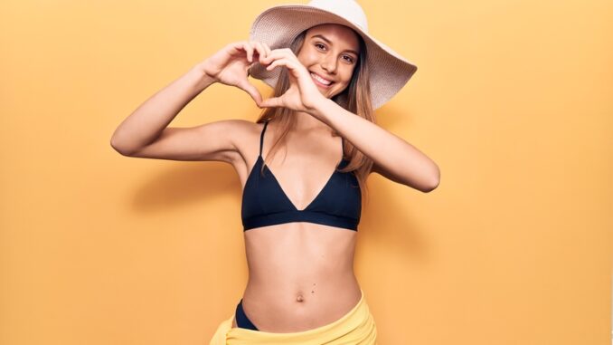 Young beautiful girl wearing bikini and hat smiling in love doing heart symbol shape with hands