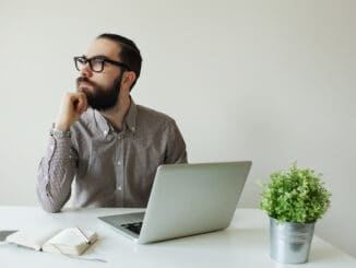 Busy man with beard in glasses thinking over laptop with smartphone on the table