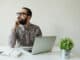 Busy man with beard in glasses thinking over laptop with smartphone on the table