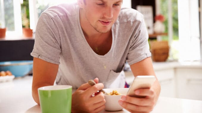 dreamstime_s_59718503 Man Eating Breakfast Whilst Checking Mobile Phone