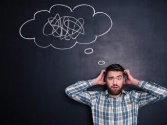 Confused bearded young man grabbing his head and thinking about problem with black board behind him