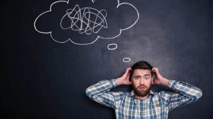 Confused bearded young man grabbing his head and thinking about problem with black board behind him