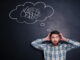 Confused bearded young man grabbing his head and thinking about problem with black board behind him