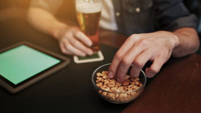 People, leisure and technology concept - close up of man with tablet pc computer drinking beer and eating peanuts at bar or pub