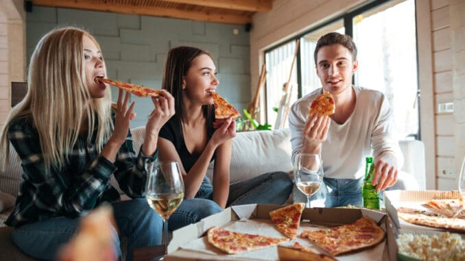 Three friends are sitting on sofa and eating pizza in house