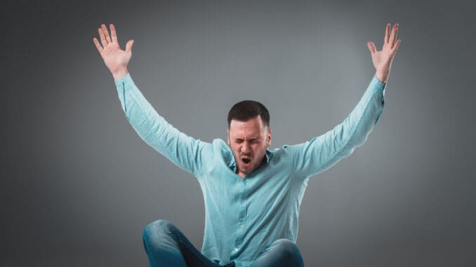 Casual young man sitting on the floor with his legs crossed and cheering with his hands in the air